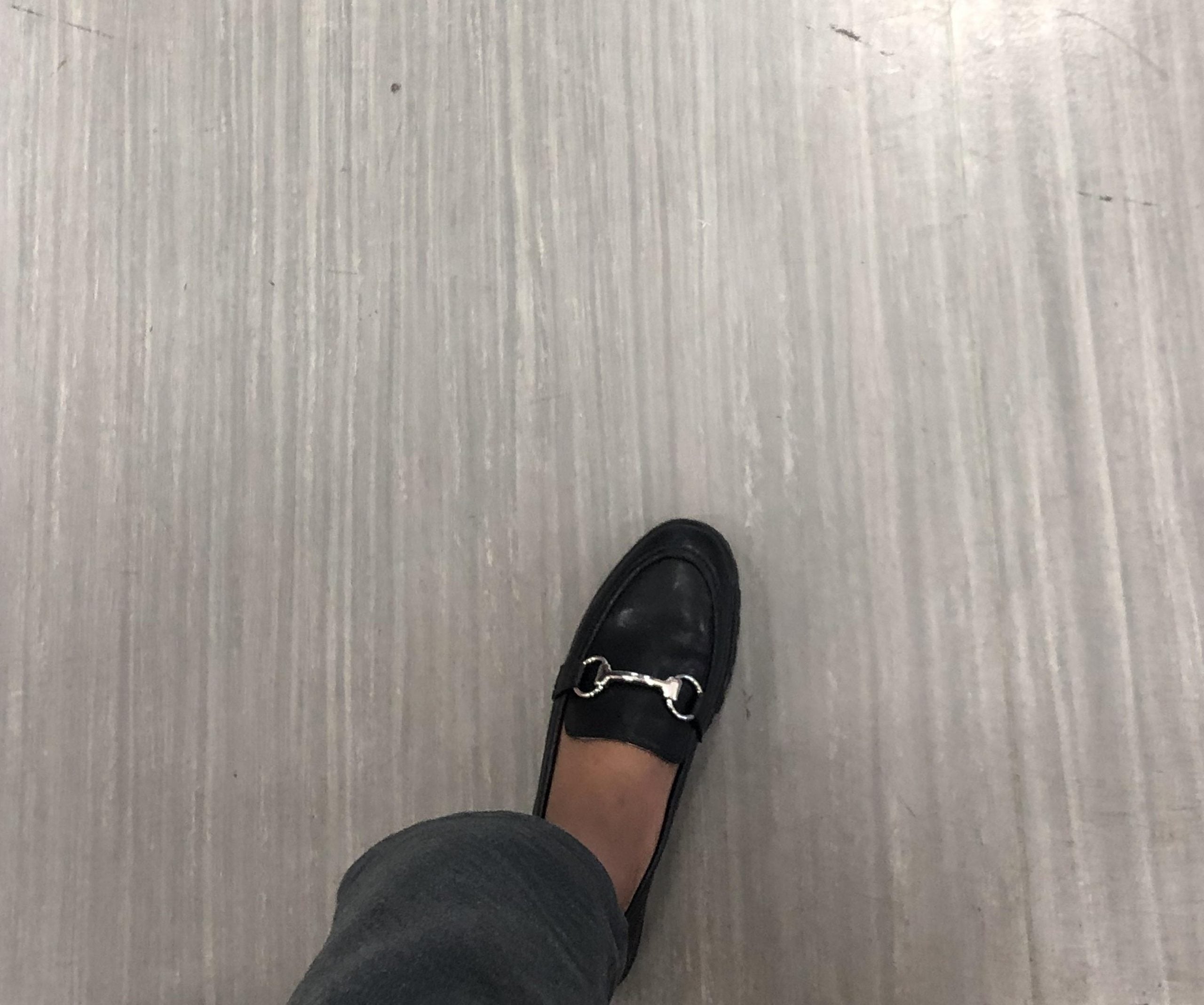 A black loafer with a silver horsebit detail worn with dark pants on a light-colored floor.
