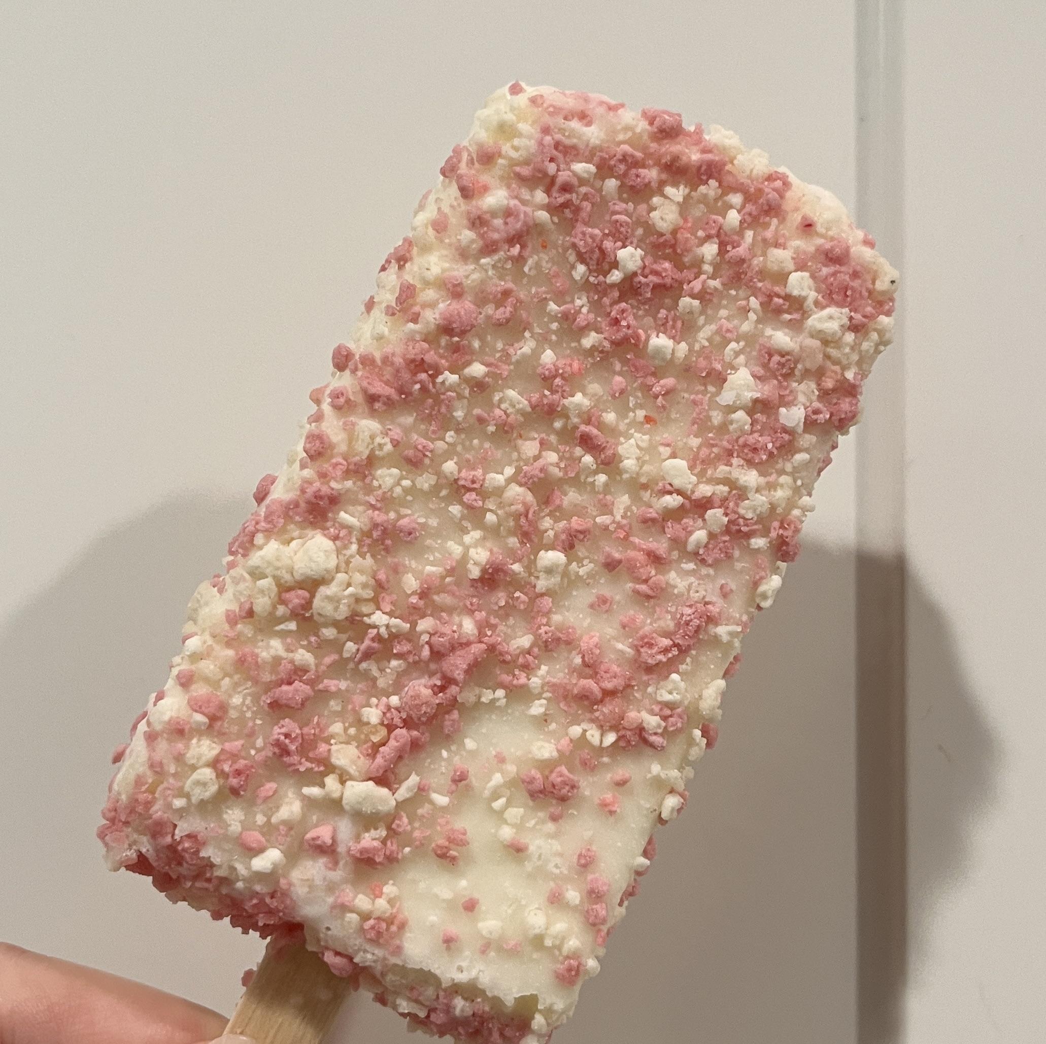 A rectangular ice cream bar coated in white icing and sprinkled with pink and white cookie crumbles on a wooden stick.