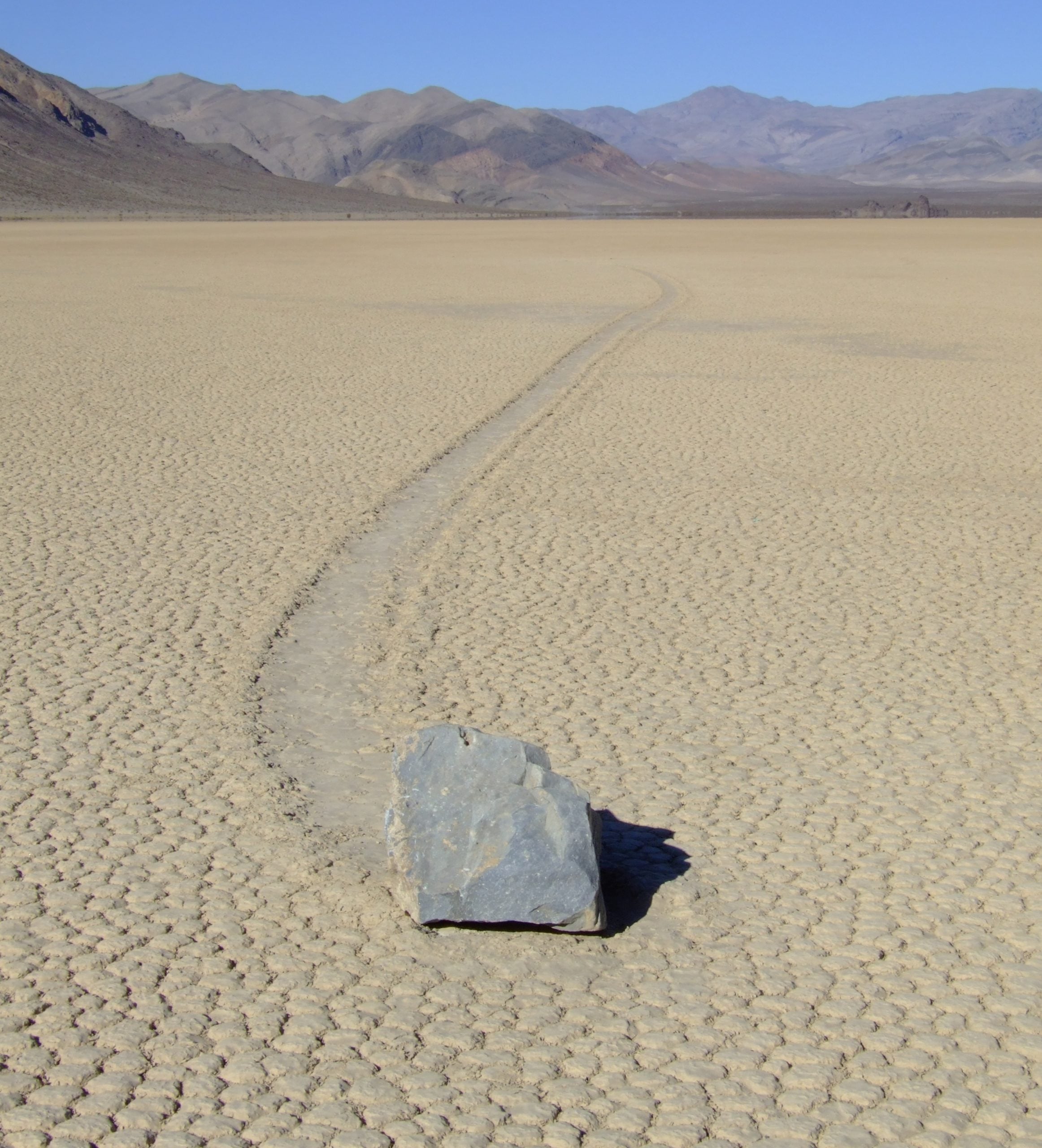 A large rock on a cracked, dry lake bed with a long trail carved into the ground behind it, leading toward distant mountains.