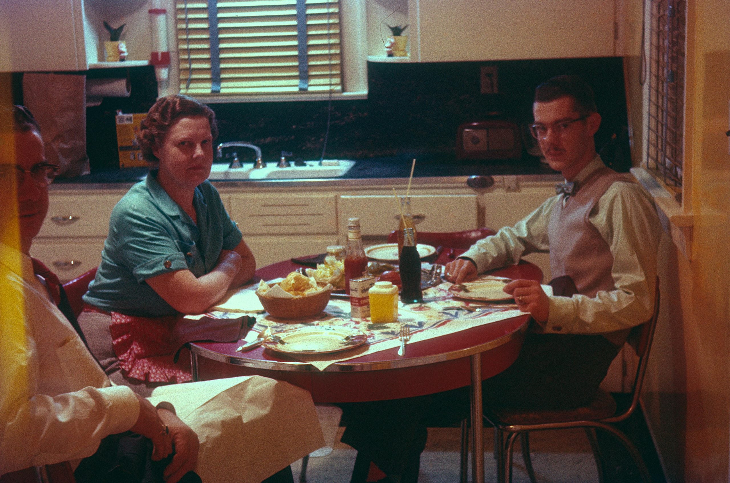 Three people sit around a round dining table set with plates, a bowl of chips, and condiments in a kitchen.