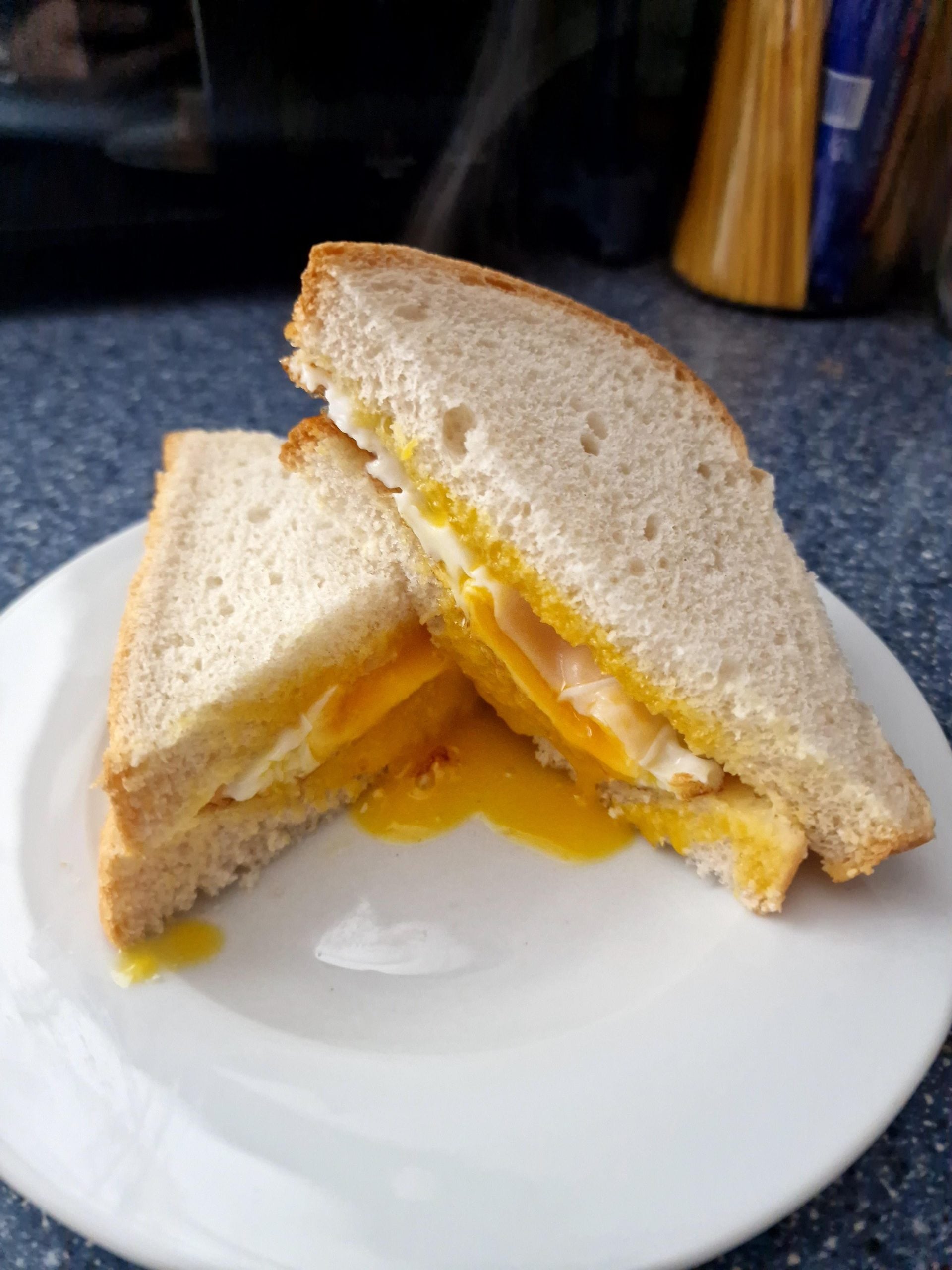 Two halves of a sandwich with egg and turkey on white bread sit on a white plate. The egg yolk is runny and dripping onto the plate. The background shows a blue countertop and kitchen items.