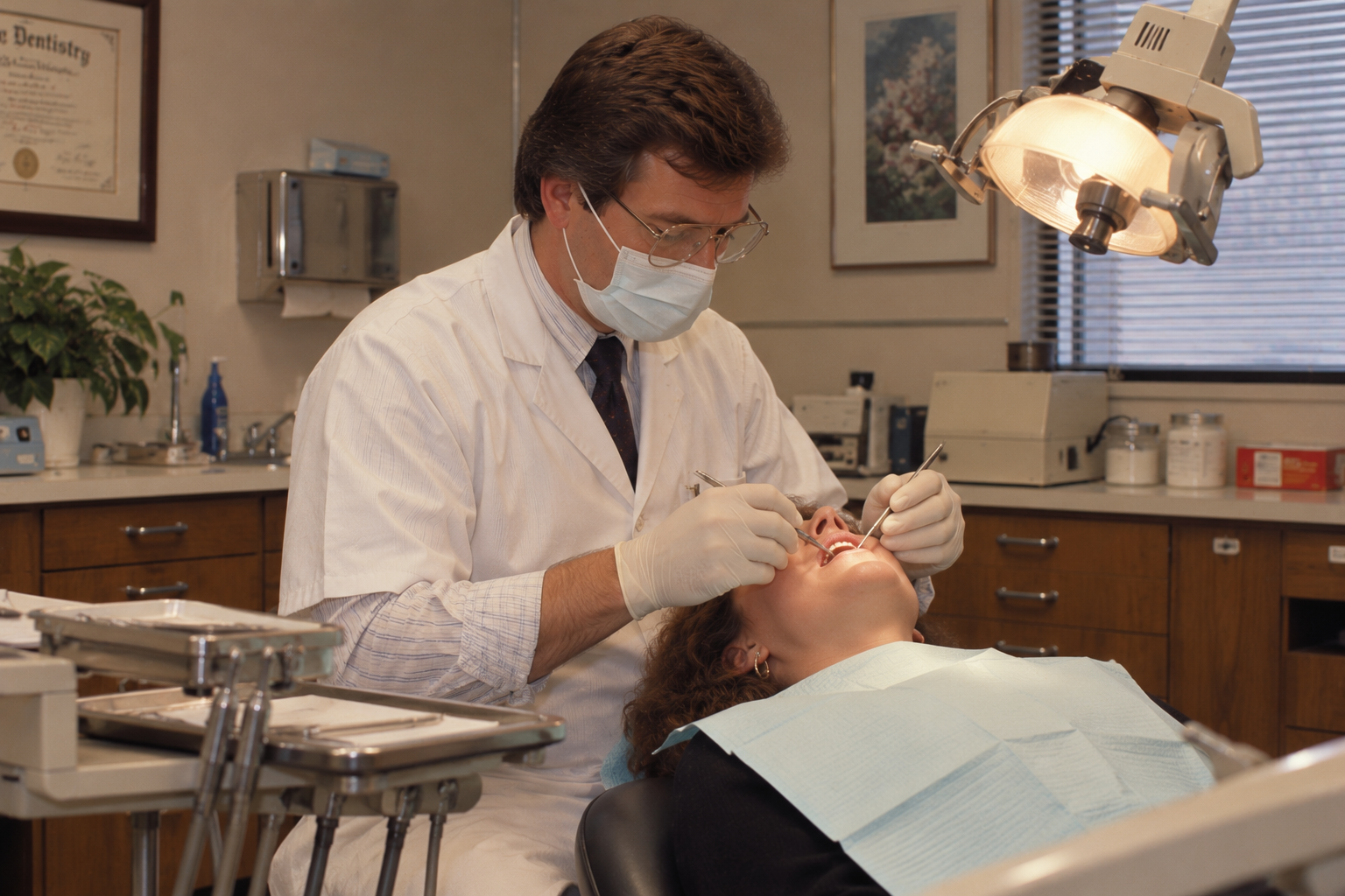 A dentist in a white coat and mask examines a patient’s teeth in a dental office.