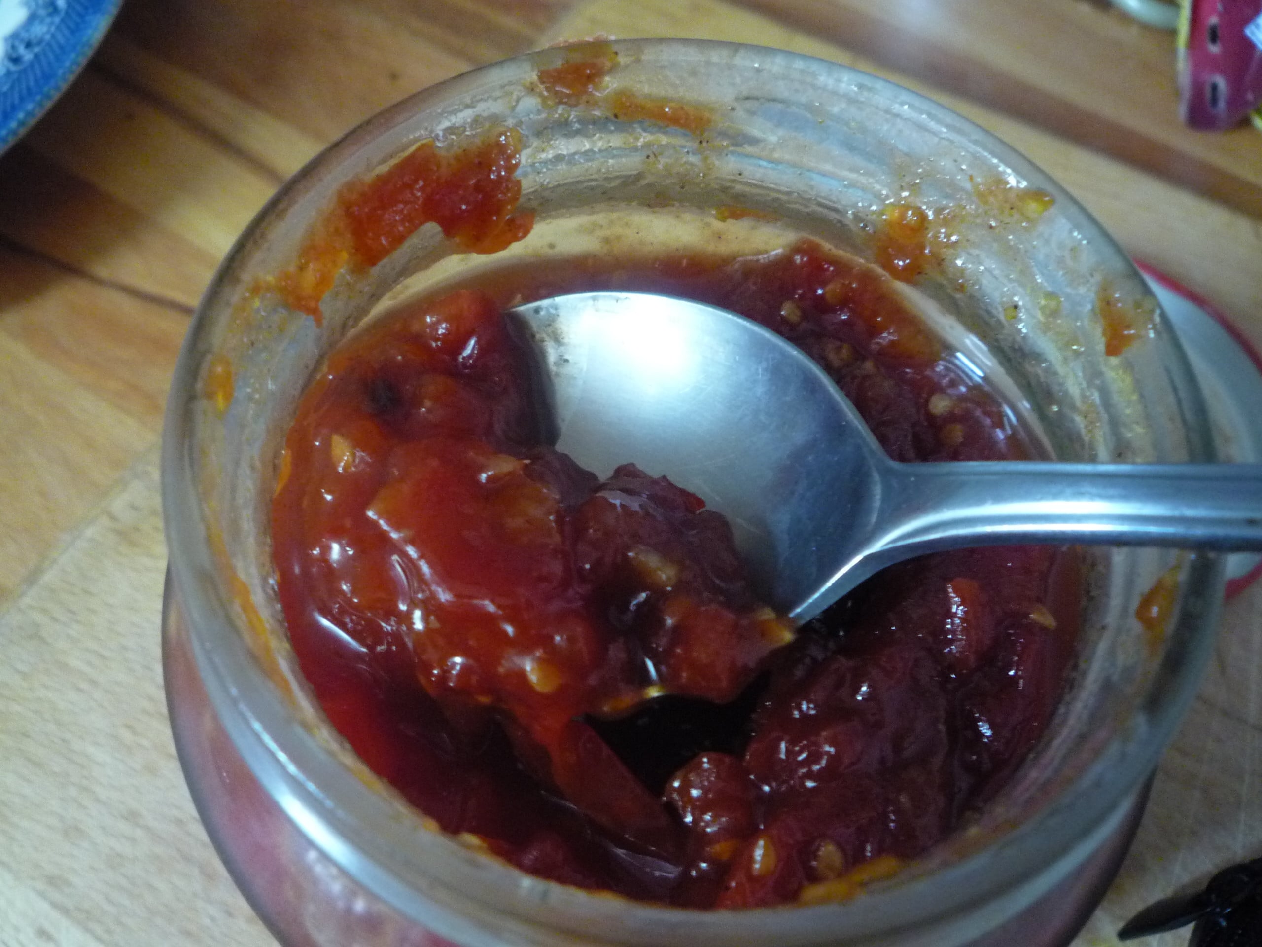A close-up of a spoon scooping chunky red tomato chutney from a glass jar, with the jar sitting on a wooden surface.