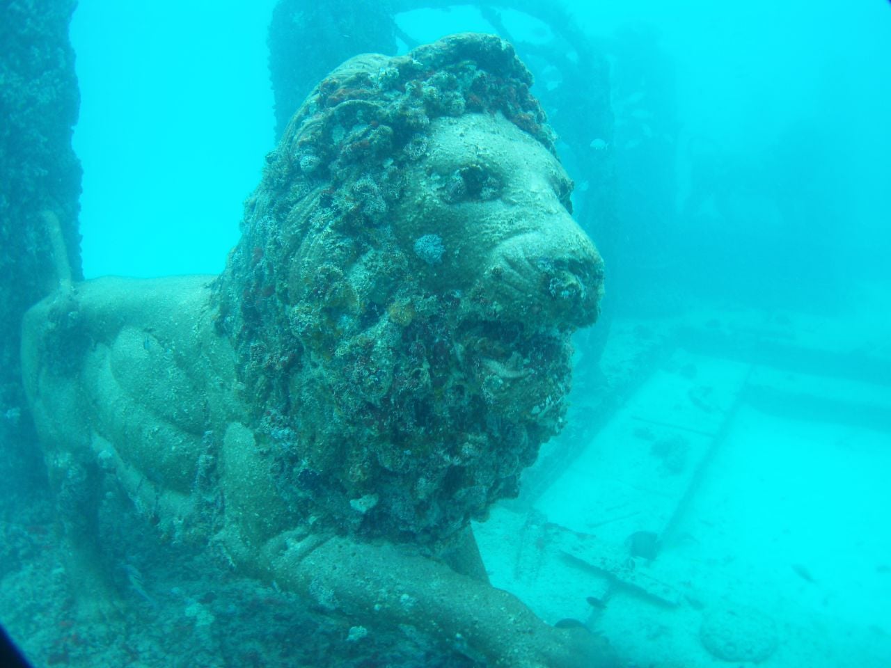 A weathered, barnacle-covered lion statue resting on the sandy ocean floor.
