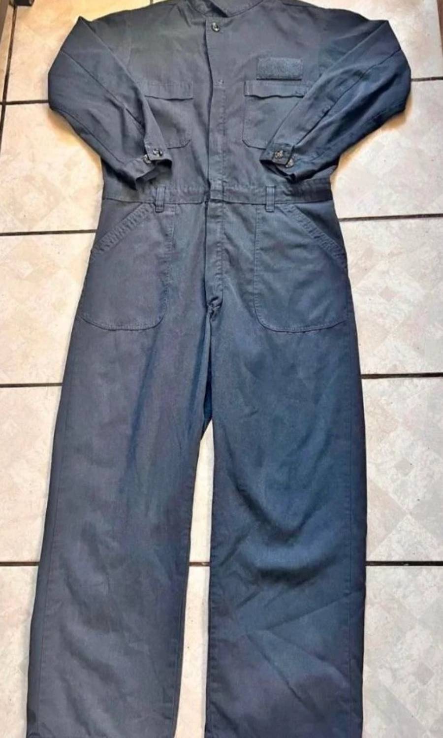 A dark blue or gray long-sleeve coverall uniform is laid flat on a tiled floor, showing buttons, pockets, and a collar.