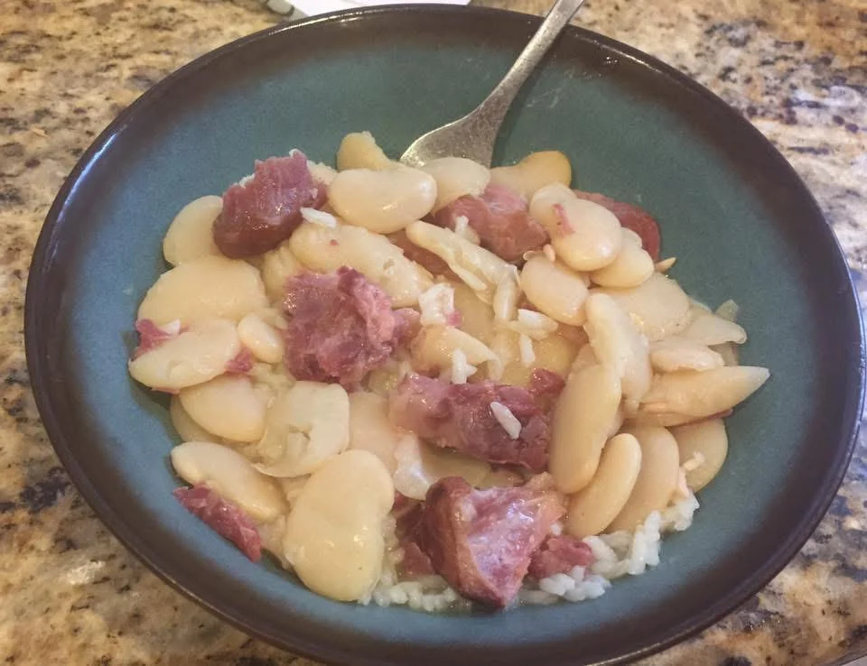 A bowl of creamy lima beans mixed with chunks of ham and sausage, served over white rice, with a fork resting in the bowl on a granite countertop.