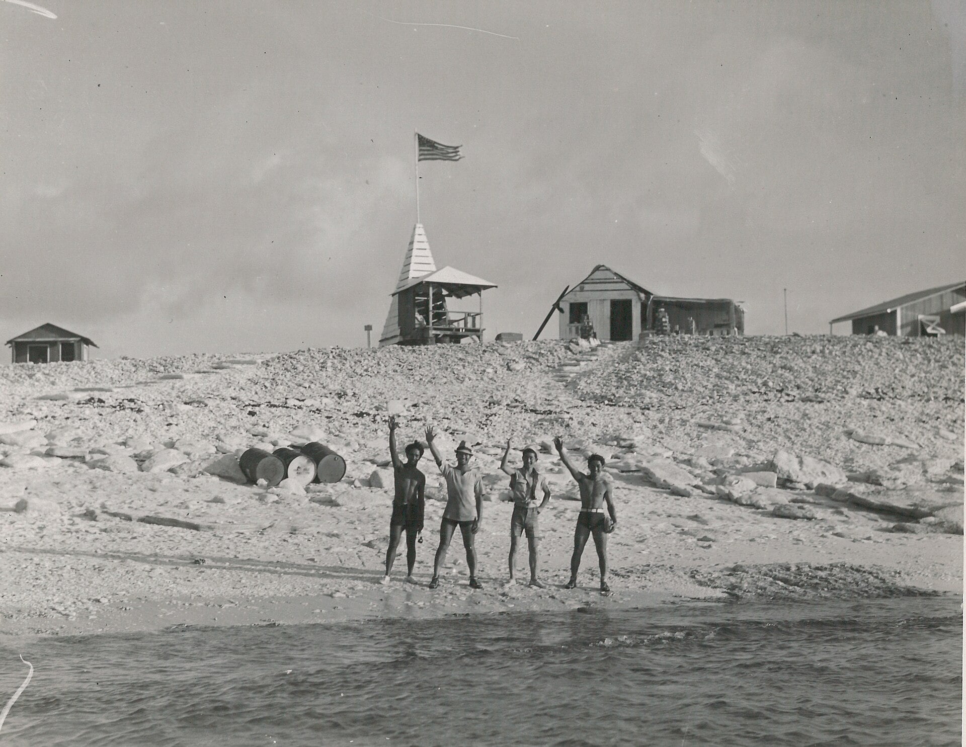 Four men stand on a rocky beach waving, with a small camp and an American flag on a pole in the background.