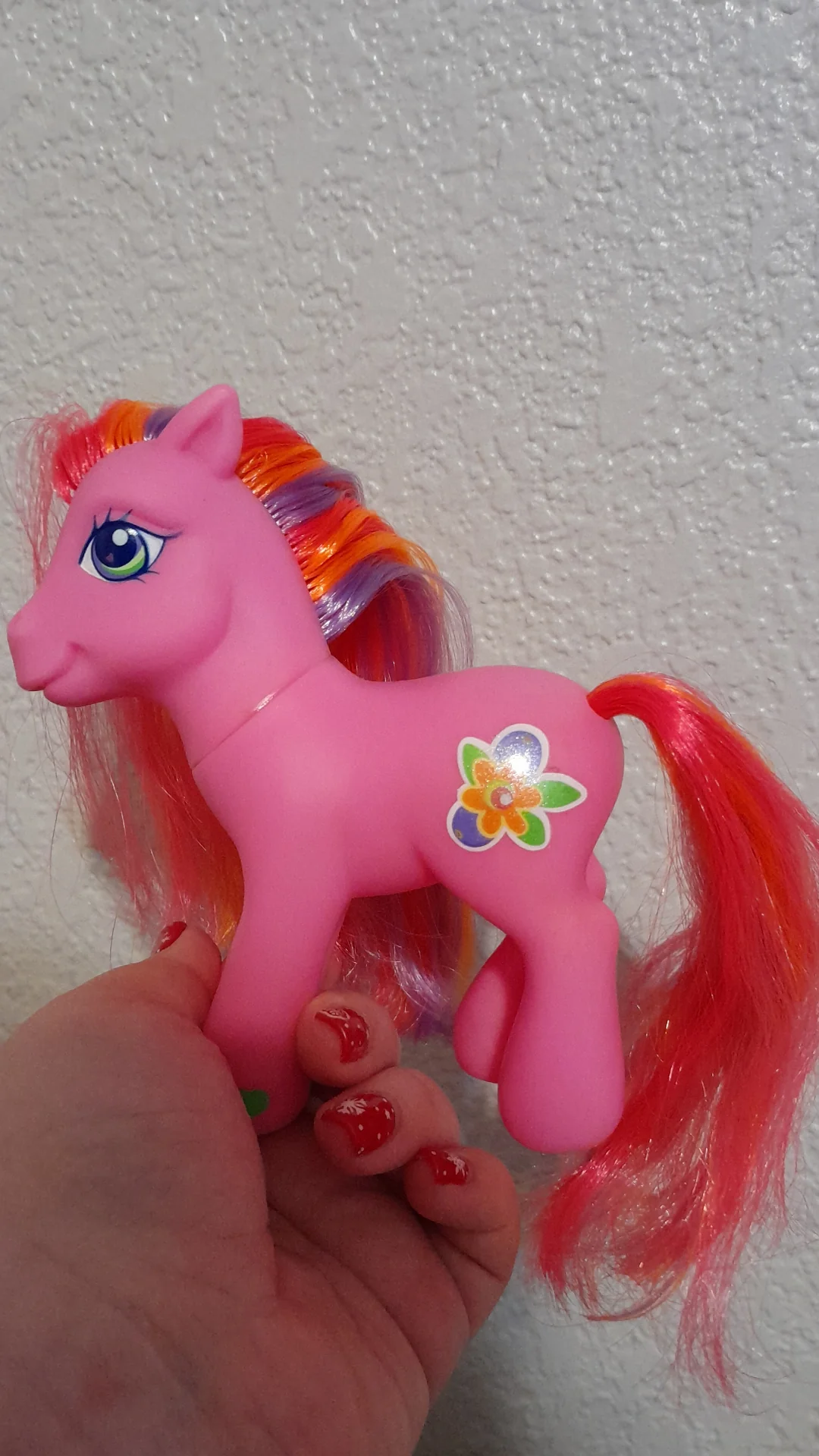 A hand holding a pink toy pony with colorful mane and tail, a green eye, and a flower symbol on its side, in front of a textured white wall.