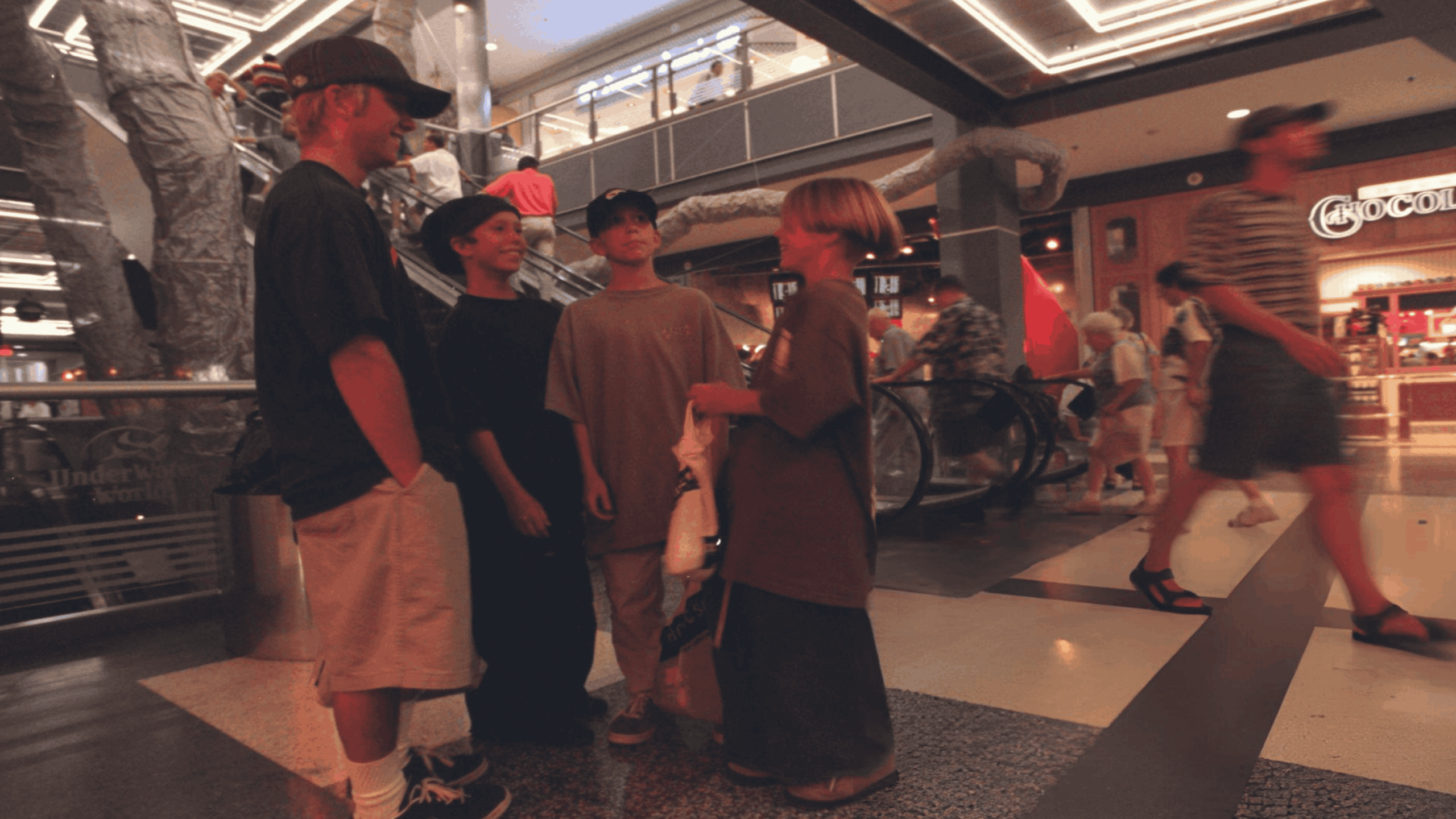 Four young people stand talking in the middle of a busy, multi-level indoor shopping mall.