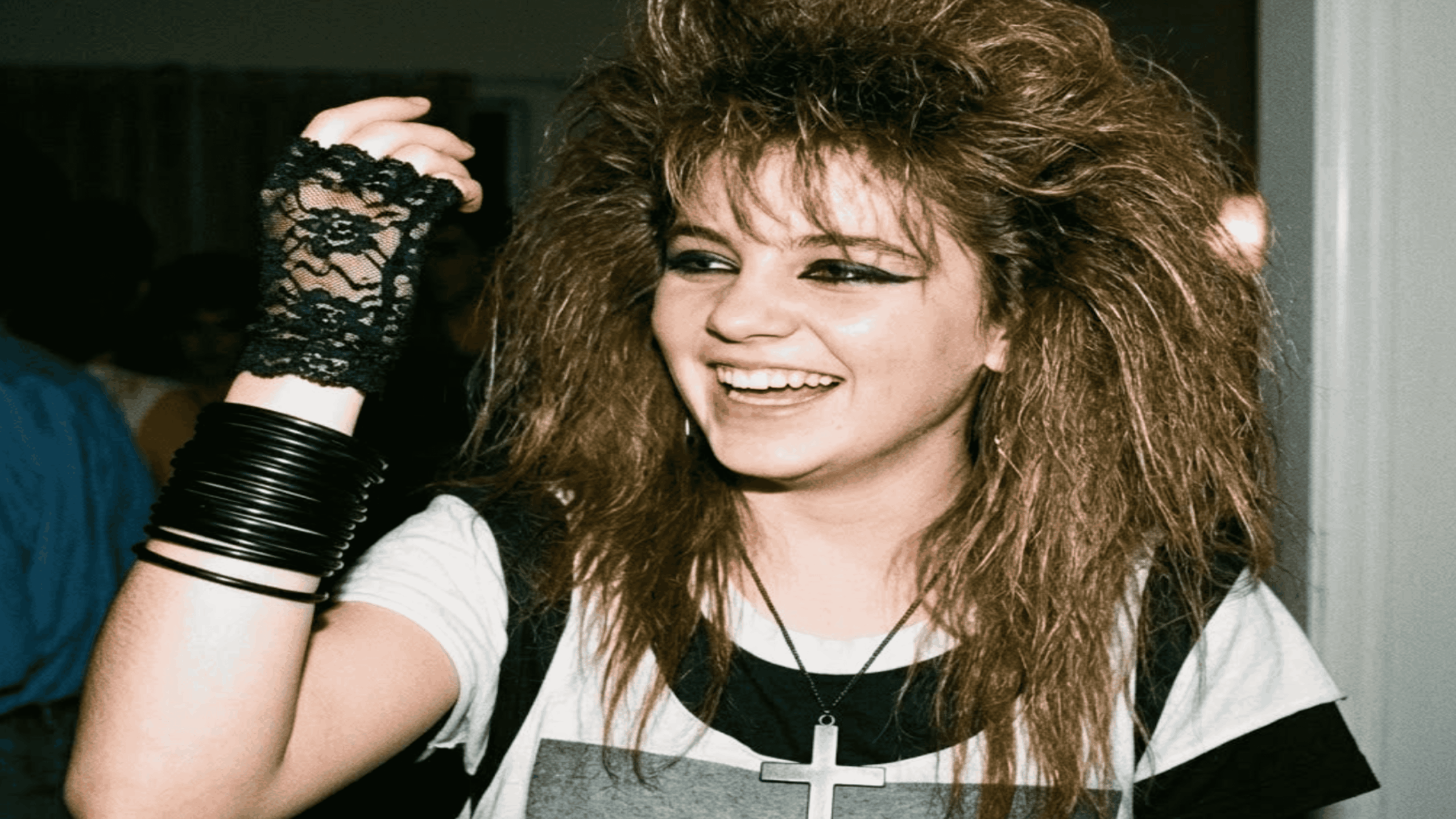 A young woman with large 1980s-style hair wears a black lace glove, stacked bangles, and a cross necklace.