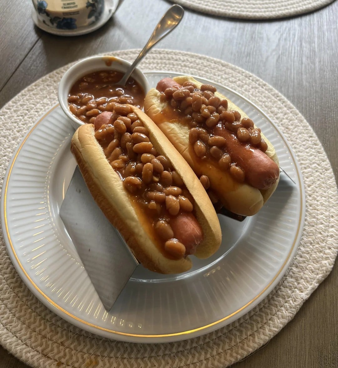 Two hot dogs in buns topped with baked beans are on a white plate. A small bowl of baked beans with a spoon sits behind them. The plate is on a woven placemat on a wooden table.