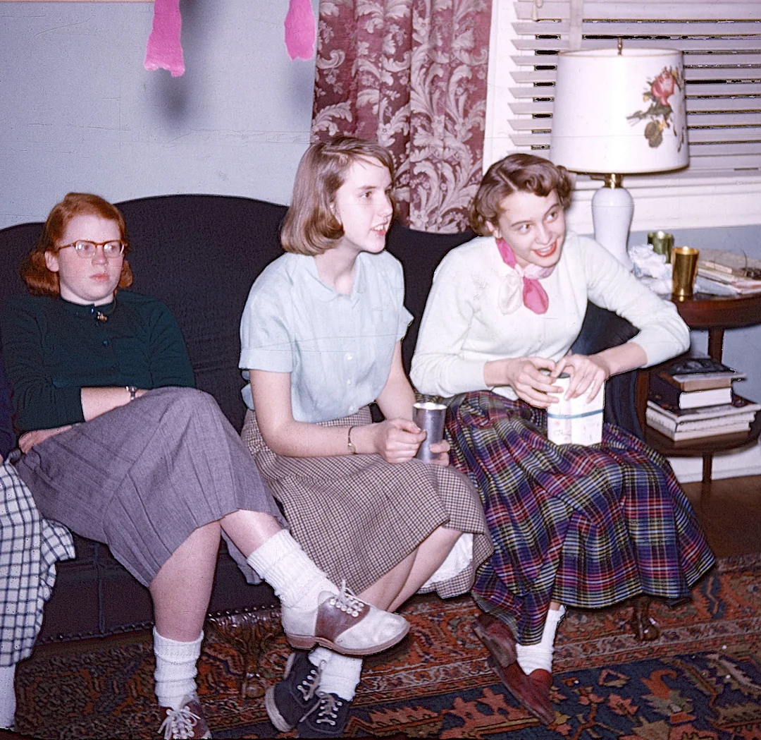 Three young women sitting on a sofa in a living room, with the woman on the right holding a small carton.