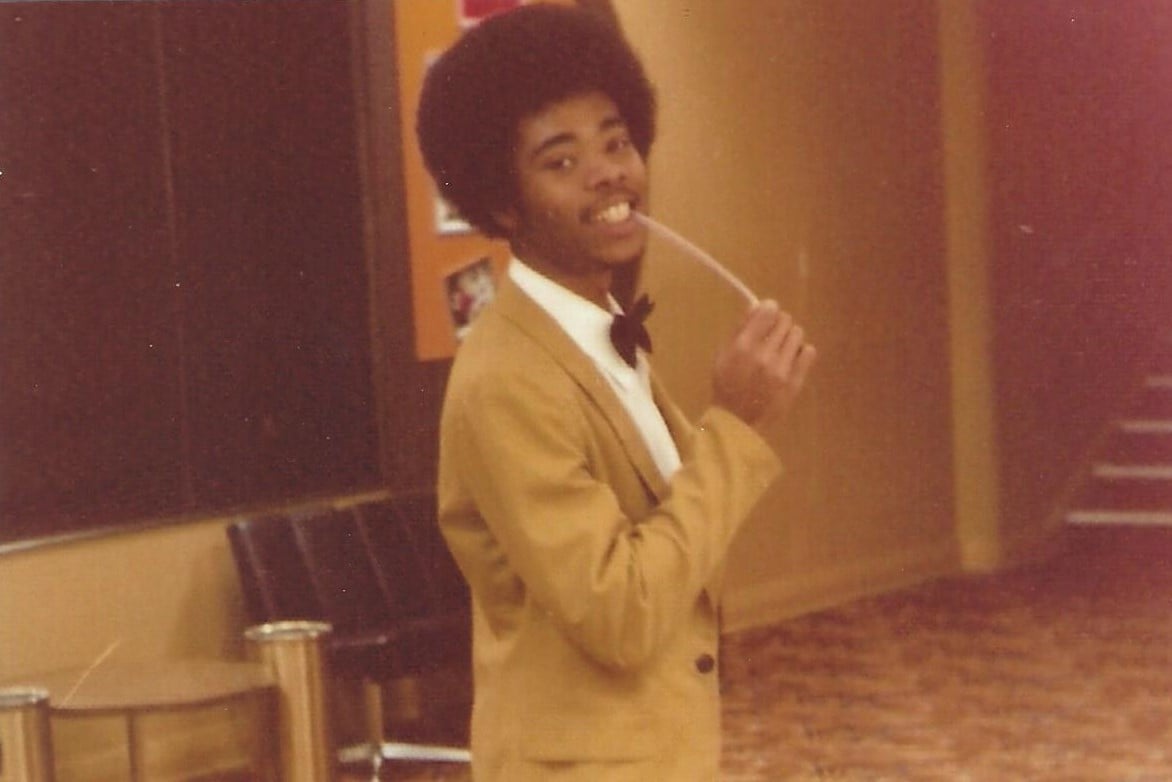 A young man with an afro wearing a tan suit and bowtie smiles while holding a straw near his mouth.