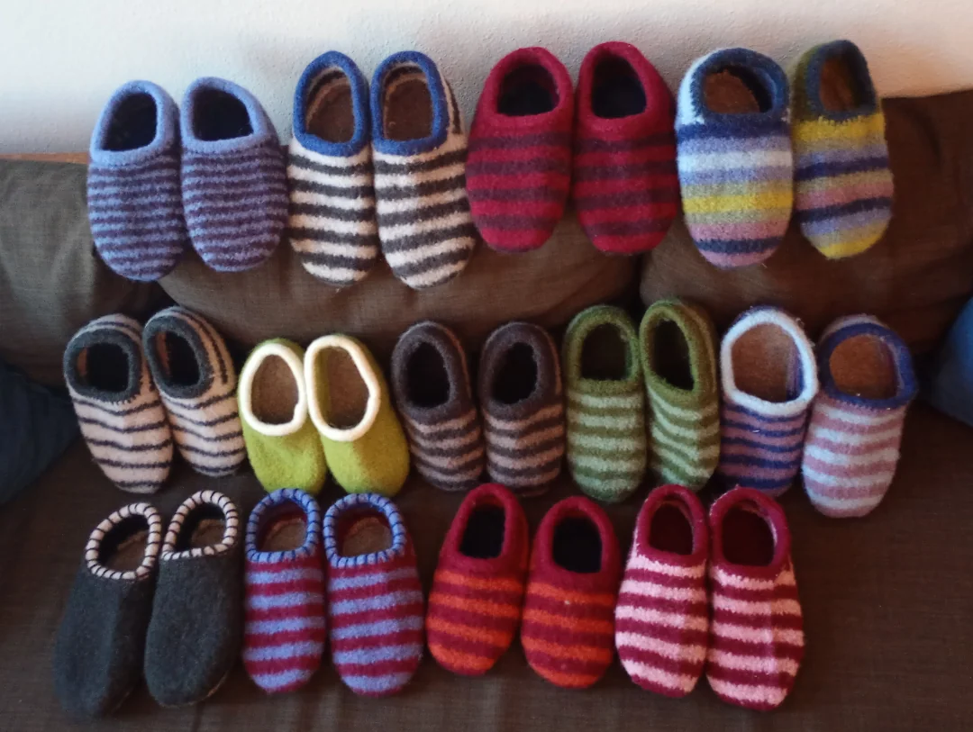 Fourteen pairs of felted wool slippers in various striped and solid colors, arranged in rows on a brown sofa.