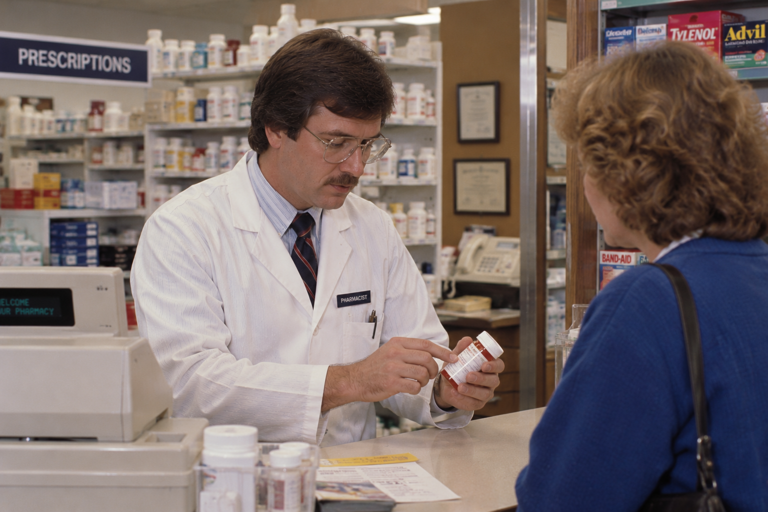 A pharmacist in a white coat explains a prescription bottle to a customer at a pharmacy counter.