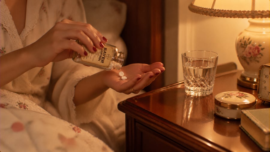 A person pouring two Bayer aspirin pills from a bottle into their hand next to a glass of water on a bedside table.