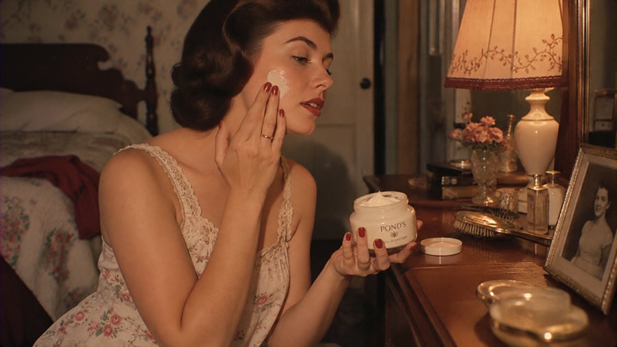 A woman in a floral camisole applying Pond’s Cold Cream to her cheek while seated at a vanity.
