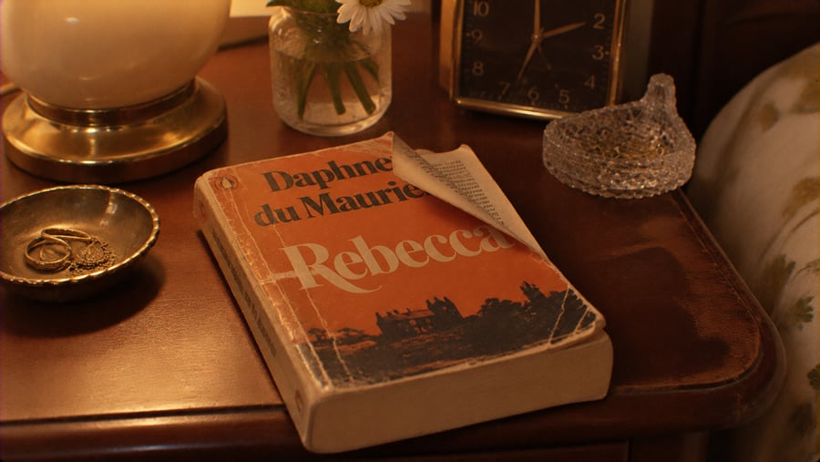 A copy of “Rebecca” by Daphne du Maurier with a dog-eared page sits on a wooden nightstand beside a lamp, a small vase of flowers, and an analog clock.