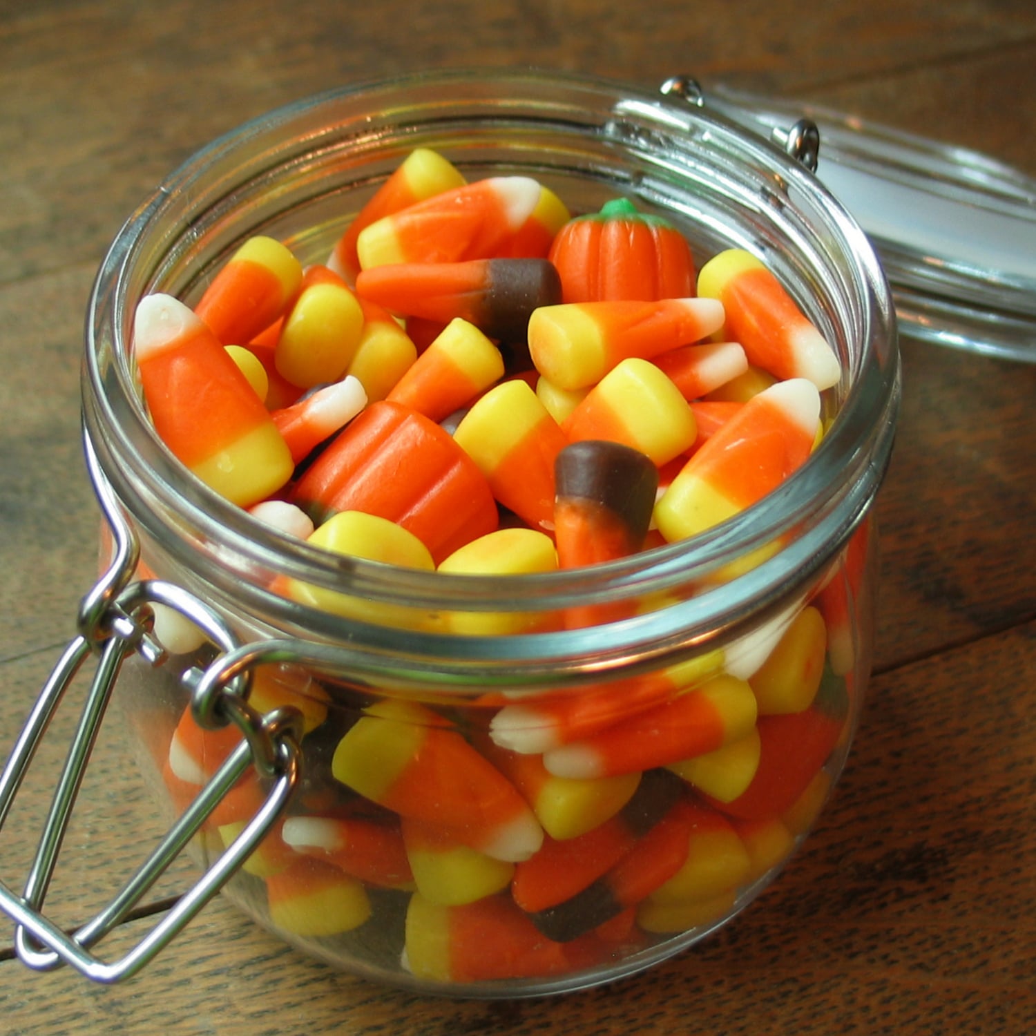 Glass jar filled with candy corn and a single candy pumpkin on a wooden surface.