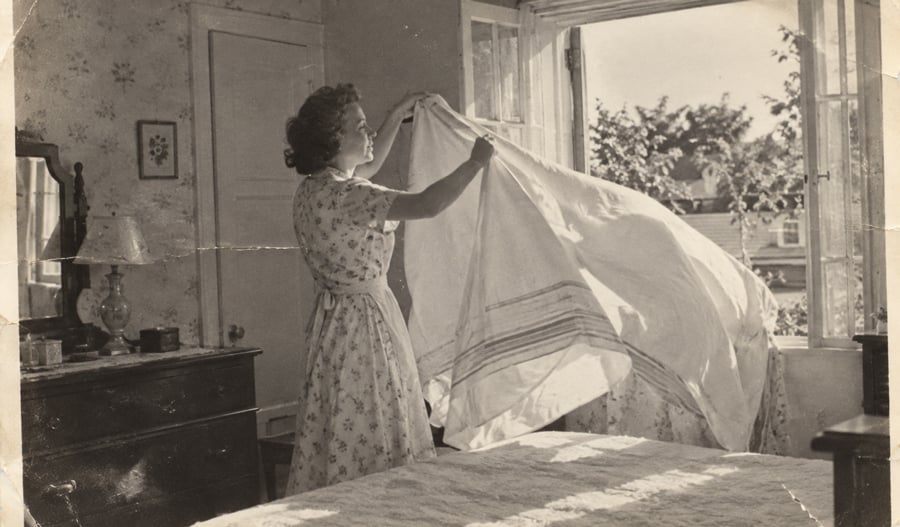 A woman in a floral dress stands by an open window, shaking out a large white sheet over a bed.