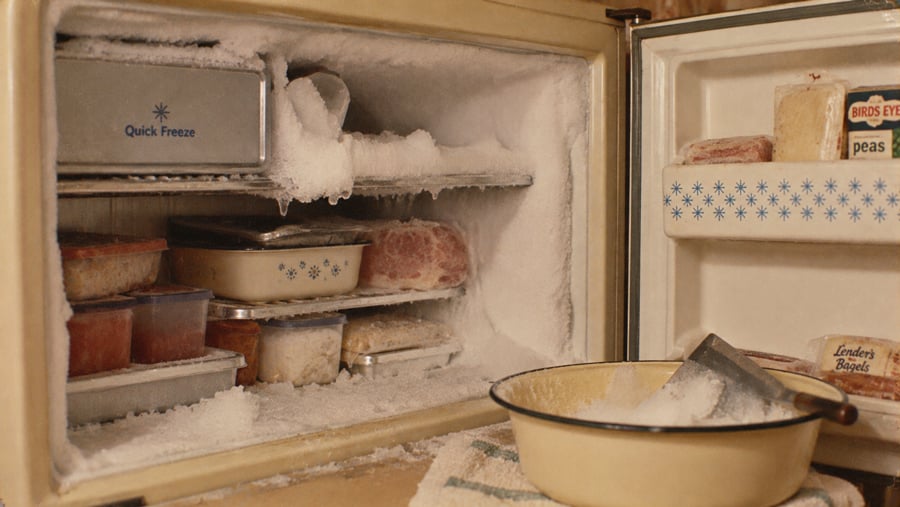 An open freezer compartment heavily coated in ice, containing various containers of food and a bowl with a scraper resting inside.