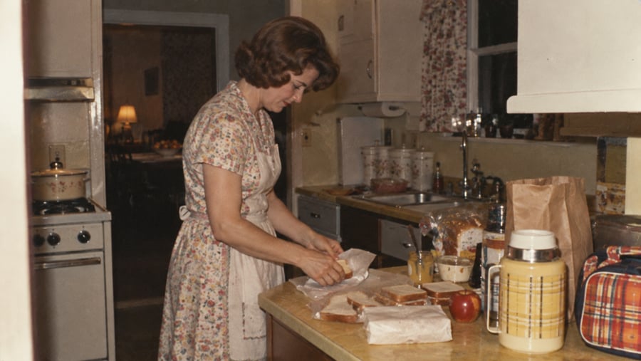A woman wearing a floral apron prepares sandwiches at a kitchen counter.