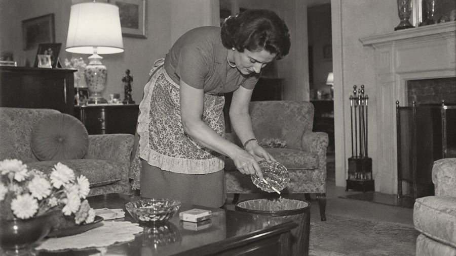 A woman wearing an apron stands in a living room, emptying a small glass bowl into a wastebasket.