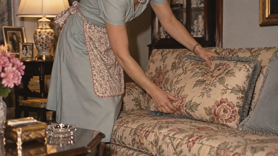 A person wearing a floral apron adjusts a matching floral throw pillow on a sofa.