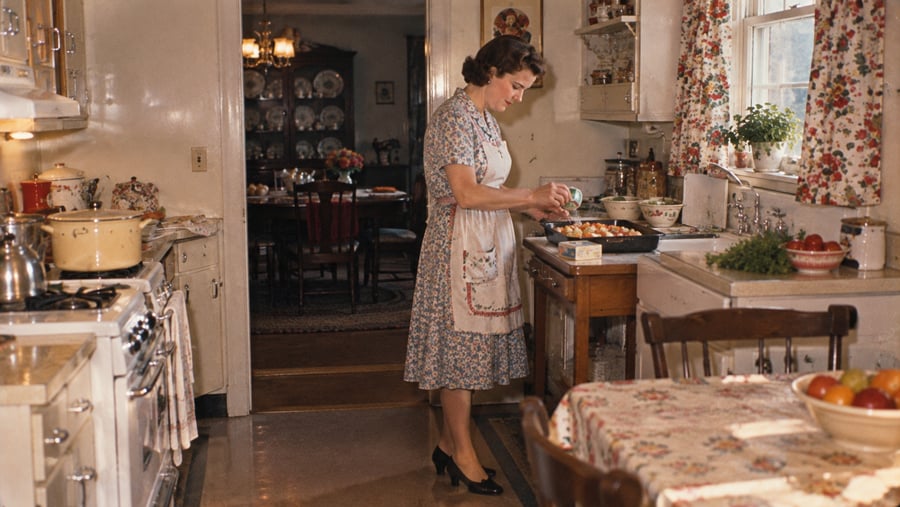 A woman in a floral dress and apron sprinkles seasoning over a baking dish in a vintage kitchen.