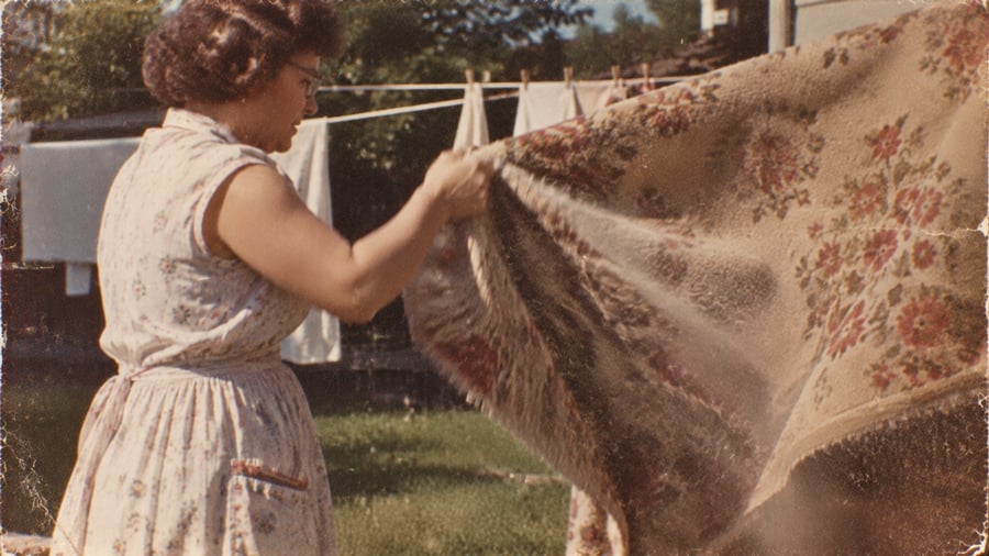 A woman in a floral dress beats dust out of a patterned rug hanging on a clothesline.