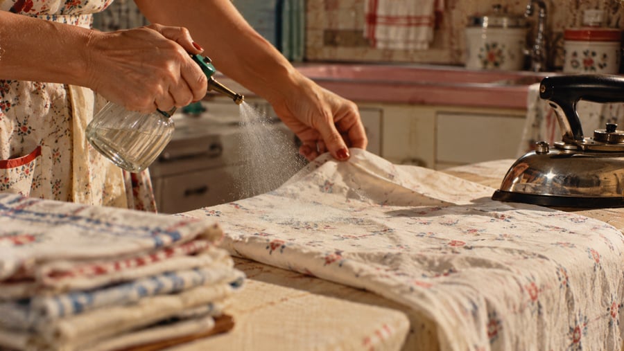 A person sprays water onto a floral patterned cloth on an ironing board, with a metal iron nearby.