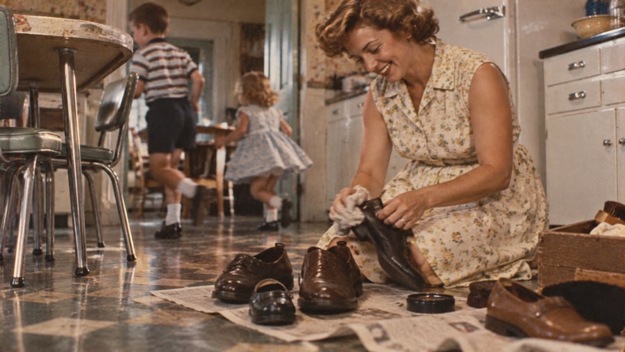 A smiling woman in a floral dress kneels on a kitchen floor, polishing leather shoes on a newspaper, while two young children play in the background.