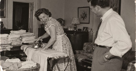 A woman in a floral apron smiles while ironing a garment on an ironing board, while a man in a dress shirt and tie watches her from across the room.