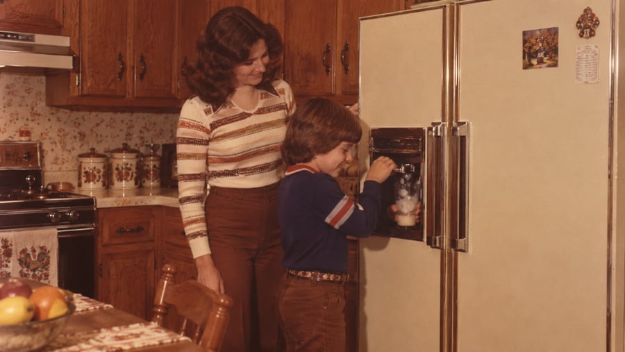 A woman watches a young boy filling a glass with ice from a refrigerator door dispenser in a kitchen.