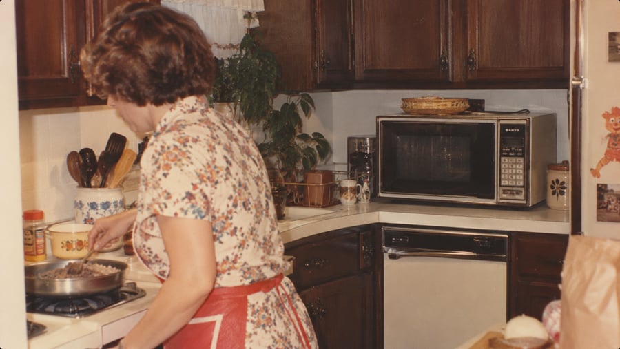 A woman in a floral apron stirs a pan on a stovetop in a kitchen.