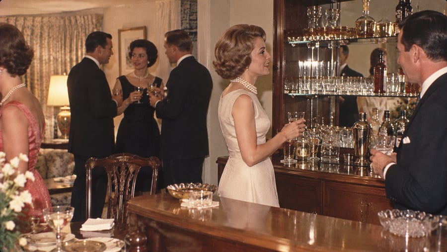 A woman in a cream dress and pearl necklace holds a drink while talking to a man at a home bar, with other couples socializing in the background.