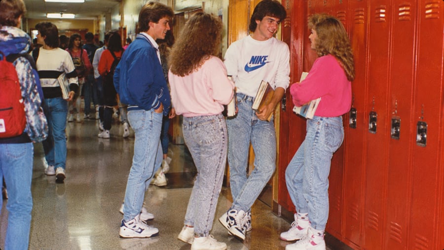 Four teenagers in 1980s fashion stand and talk in a school hallway lined with red lockers.