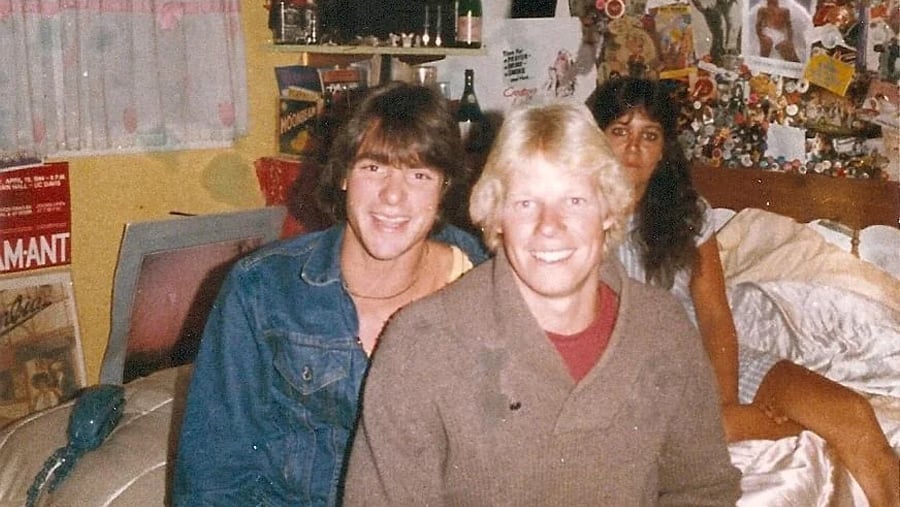 Two young men and a young woman sit together on a bed in a room with posters on the walls.