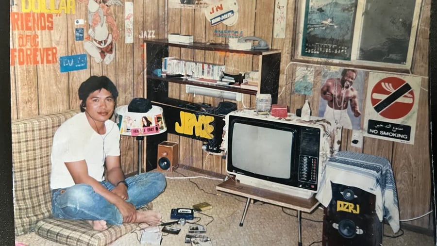 A man sitting on a floor cushion in a room with wood-paneled walls, a vintage television, cassette tapes, and various posters.