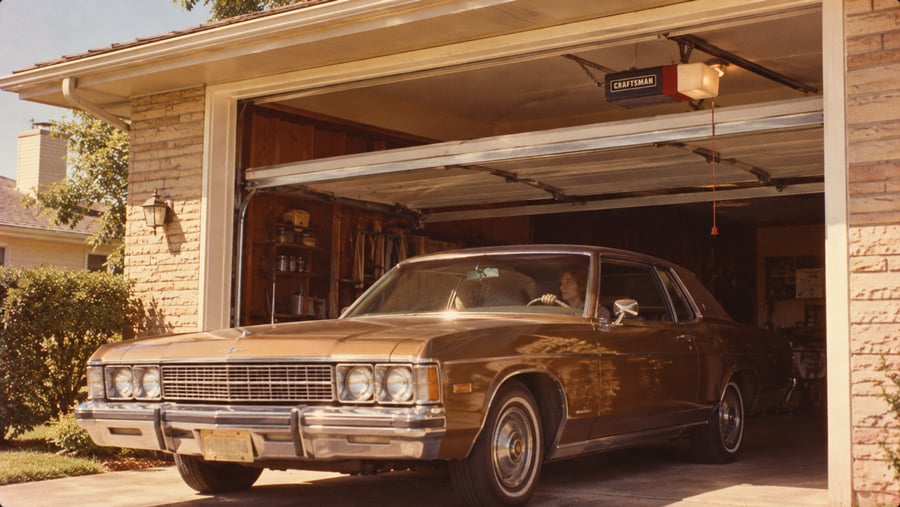 A person sits in the driver’s seat of a brown vintage sedan parked inside a residential garage.