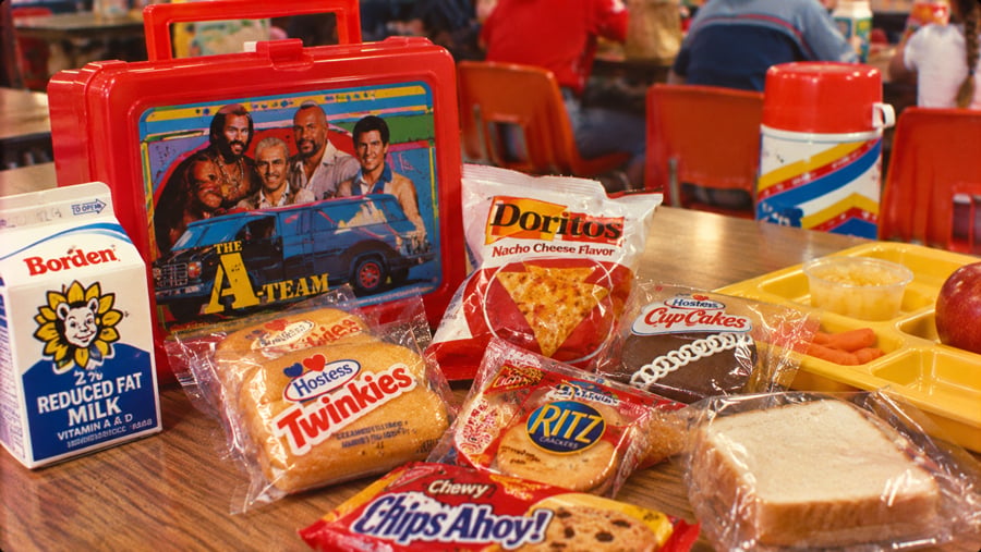 An A-Team lunchbox on a cafeteria table surrounded by various snacks, including a carton of Borden milk, Twinkies, Doritos, Ritz crackers, a Hostess cupcake, a sandwich, and a thermos.