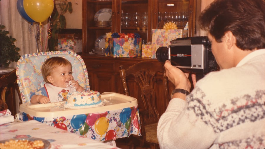A person holding a vintage RCA video camera films a toddler in a high chair eating a birthday cake.