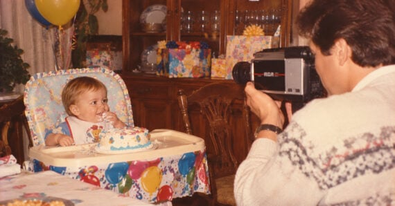 A man filming a baby in a high chair eating a birthday cake with a vintage RCA camcorder.