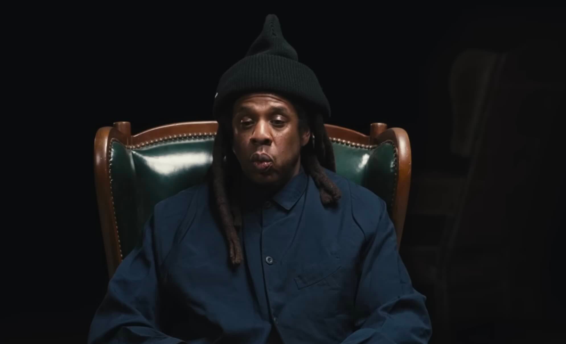Jay-Z wearing a black pointed beanie and a navy blue button-down shirt, sitting in a green leather chair against a black background.