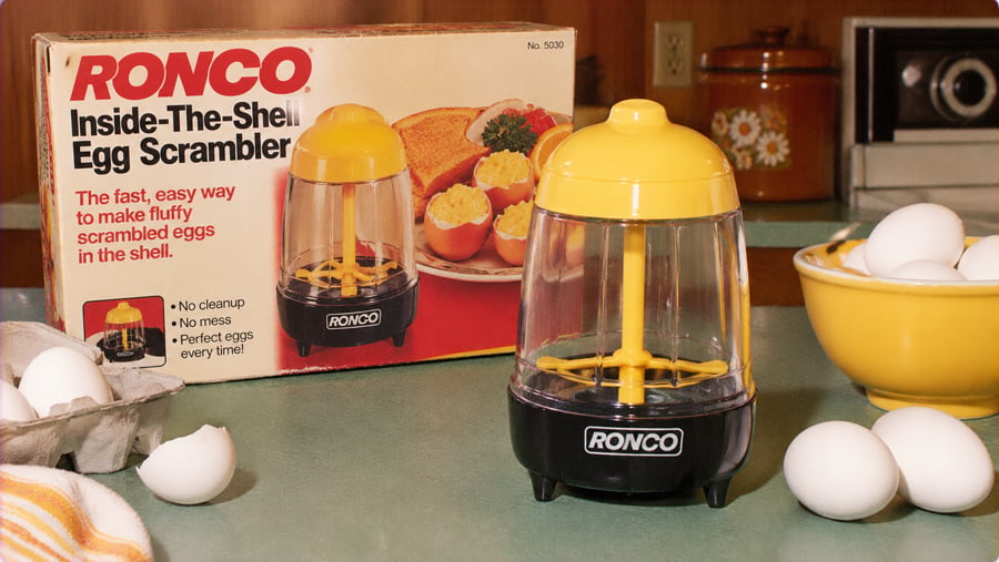 A Ronco Inside-The-Shell Egg Scrambler device sits on a kitchen counter next to its retail box and a bowl of raw eggs.