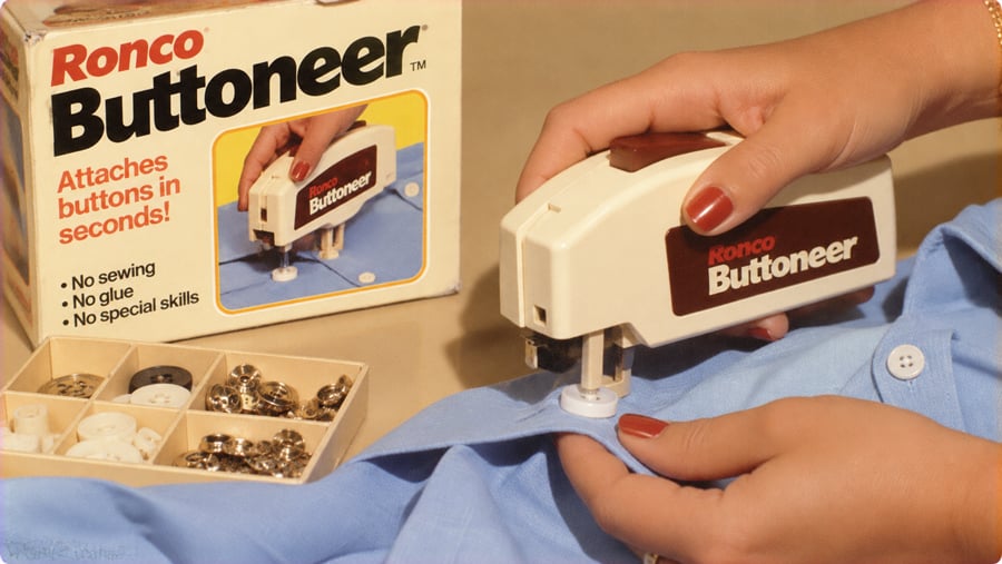 A hand uses a Ronco Buttoneer handheld tool to attach a button to a light blue shirt, with the product packaging and a box of spare buttons nearby.