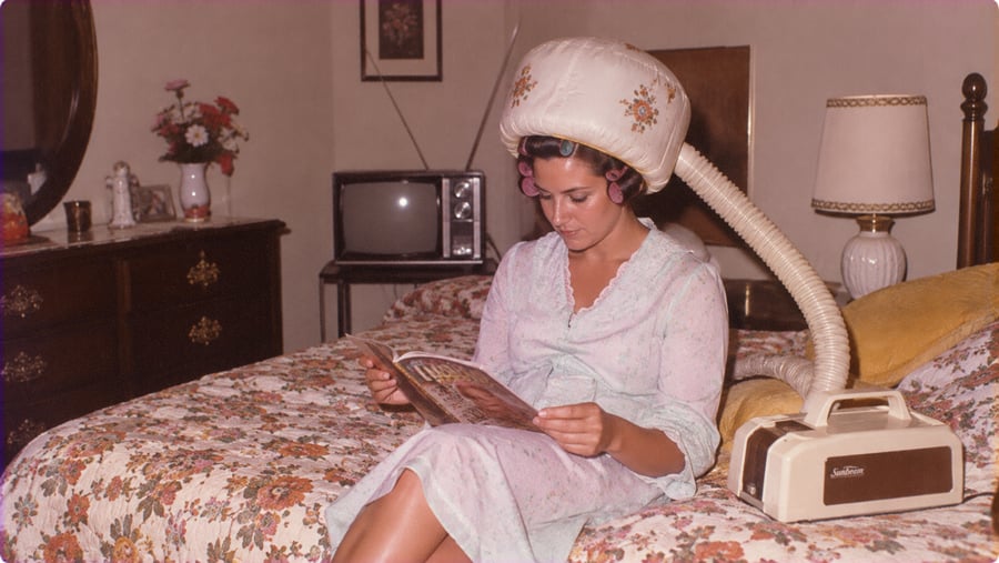 A woman in a floral nightgown and hair curlers sits on a bed reading a magazine while wearing a portable hooded hair dryer.