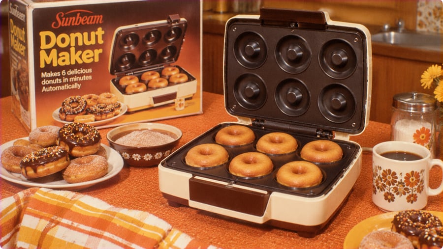 A Sunbeam donut maker with its lid open, revealing six baked donuts, next to its product box and plates of finished donuts on a table.