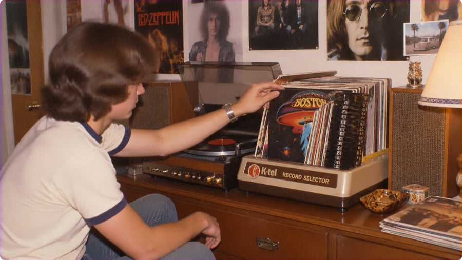 A young person browsing through a collection of vinyl records in a K-tel record selector, with posters of musicians on the wall behind them.
