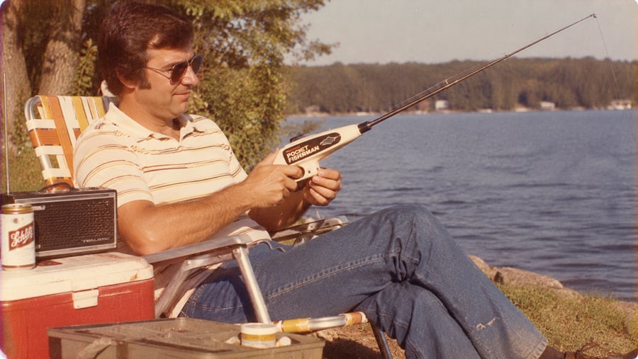 A man in a striped polo and sunglasses sits in a lawn chair by a lake, holding a Pocket Fisherman fishing rod next to a cooler with a beer and a radio.